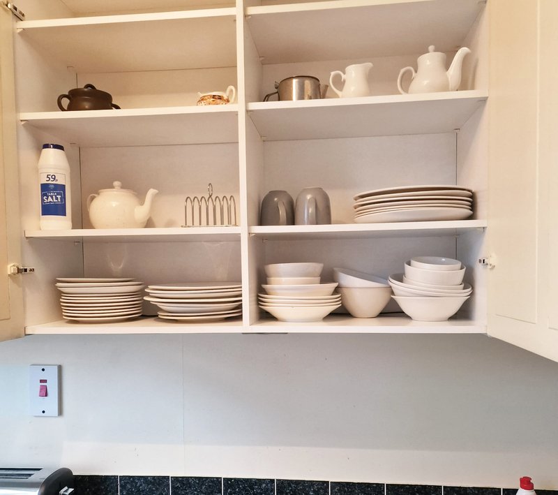 Open kitchen cupboard with white dishes, teapots, salt container, and serving utensils neatly arranged on shelves