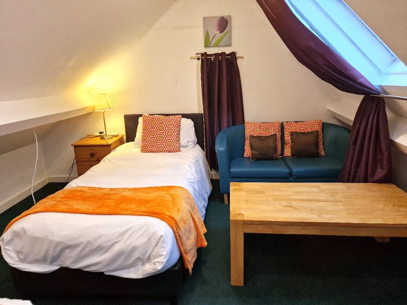 Attic bedroom with single bed, orange bedding, blue sofa with cushions, wooden table, and skylights