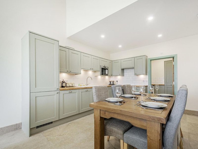Modern kitchen with sage-green cabinetry, wooden dining table set for two, and integrated appliances