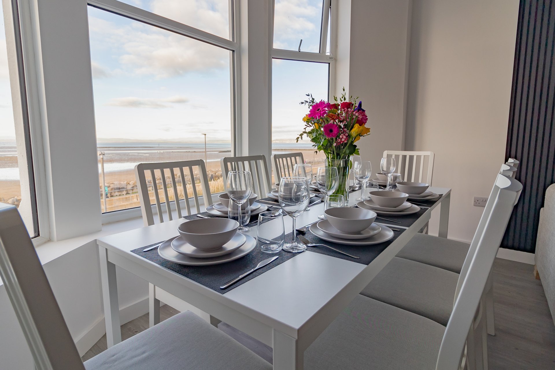 Bright dining room with set table, flowers, and coastal view through large windows