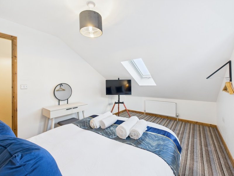 Bright attic bedroom with double bed, blue bedding, desk, TV on stand, and skylight window