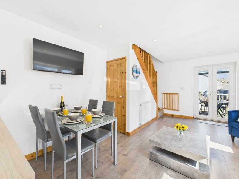 Open-plan living and dining area with dining table, grey chairs, TV, wooden doors and bright windows