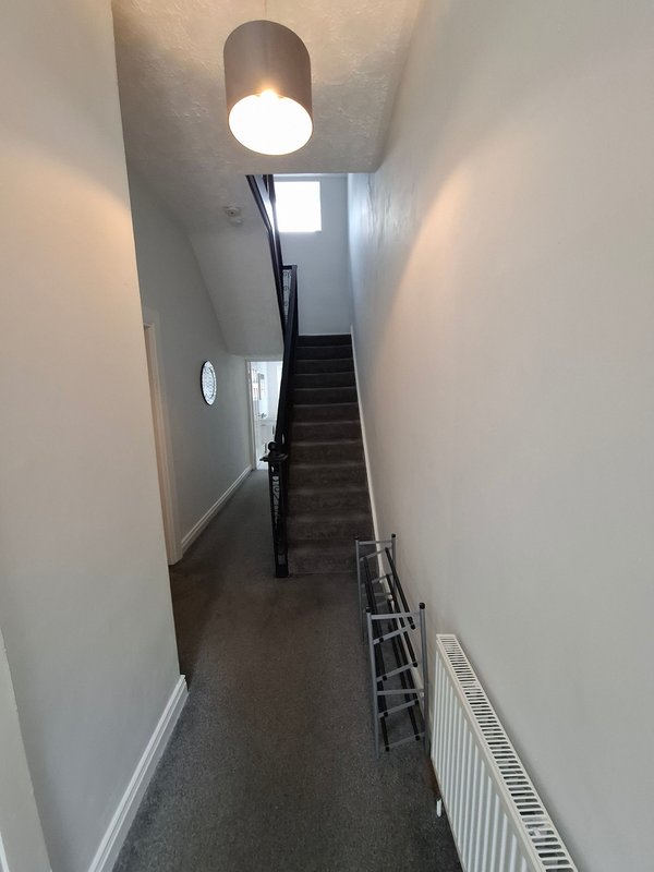 Narrow hallway with staircase, ceiling light, white walls and dark carpet flooring
