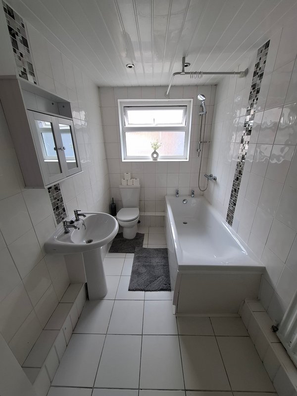Clean white-tiled bathroom with toilet, pedestal sink, bathtub and window