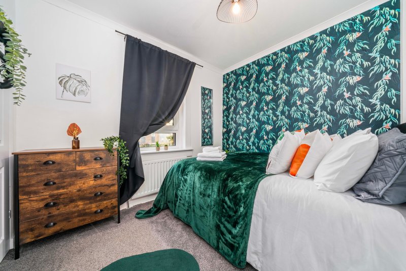 Bedroom with double bed, green velvet throw, wooden dresser, patterned wallpaper and curtains