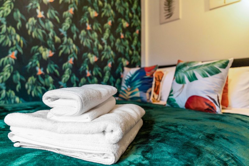 Bedroom with green bedding, rolled white towels, and tropical-patterned cushions