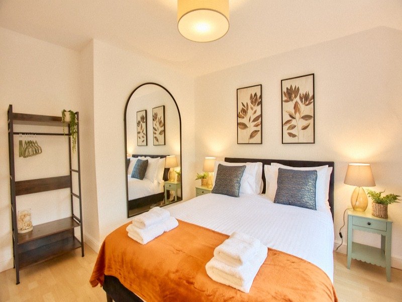 Bright bedroom with double bed, orange throw, wall mirror, framed botanical prints and wooden ladder shelf