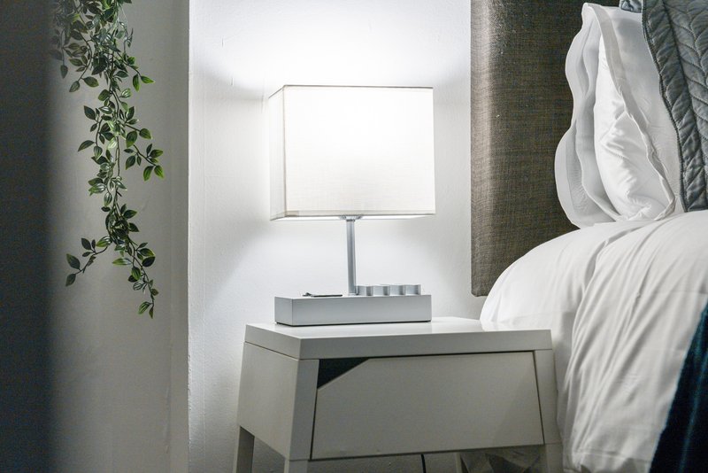 Bedroom bedside table with lamp, plant, and white bedding visible