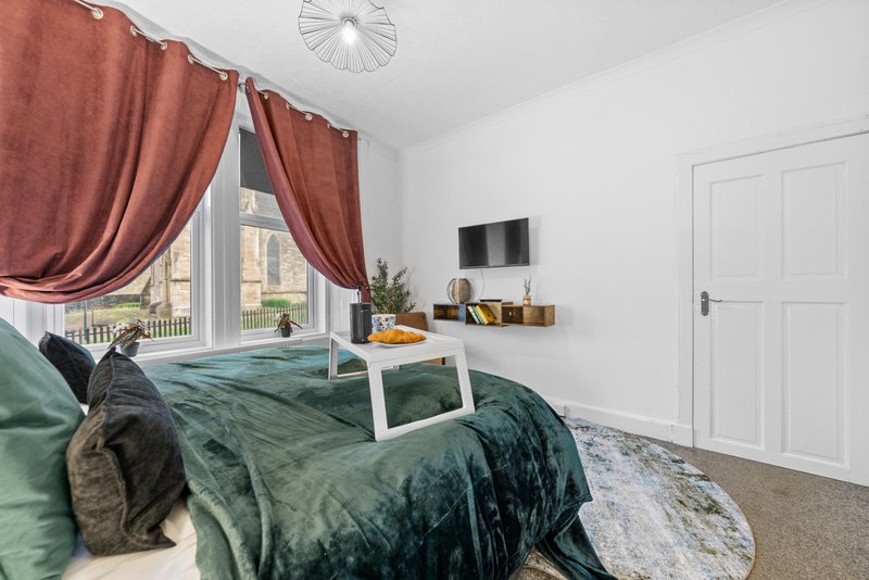 Bright bedroom with double bed, green bedding, burgundy curtains, wall-mounted TV and white door