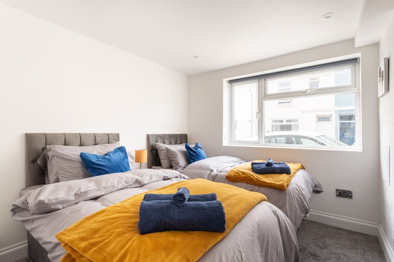 Bright modern bedroom with two beds, gray upholstered headboards, blue and yellow pillows, and large window overlooking street