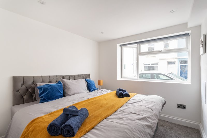 Modern bedroom with double bed, blue pillows, yellow throw, and large window with street view