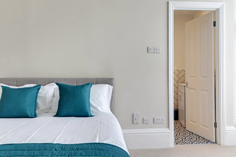 Modern bedroom with teal cushions, white bedding, grey headboard, and open door to ensuite