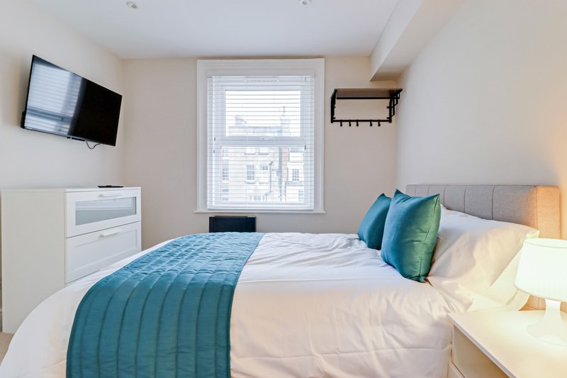 Bright bedroom with double bed, teal bedding, wall-mounted TV, and window with blinds