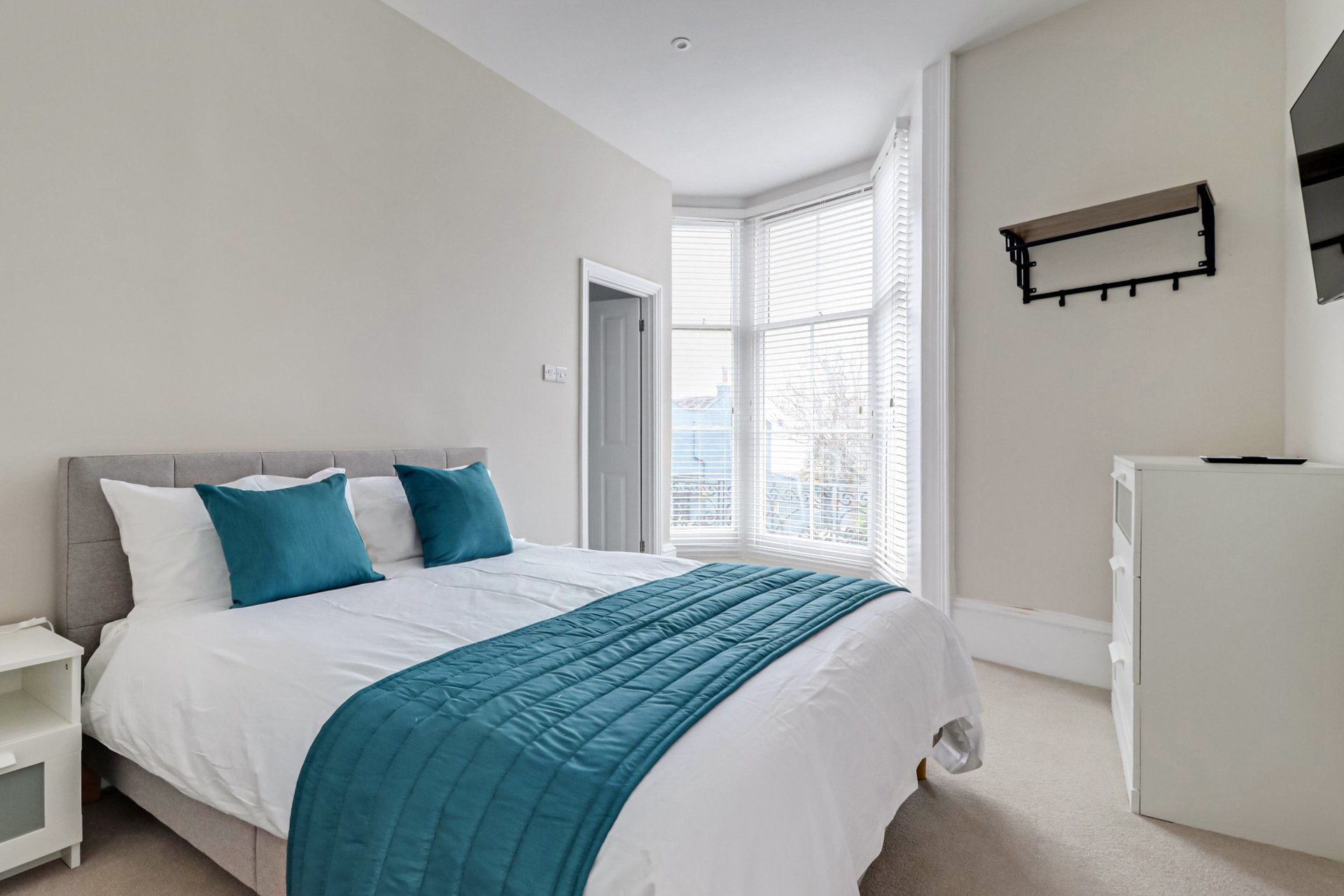 Bright modern bedroom with double bed, teal bedding, large window and white walls