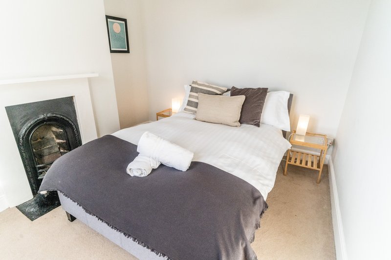 Bedroom with double bed, white bedding, dark throw blanket, fireplace and wooden bedside table