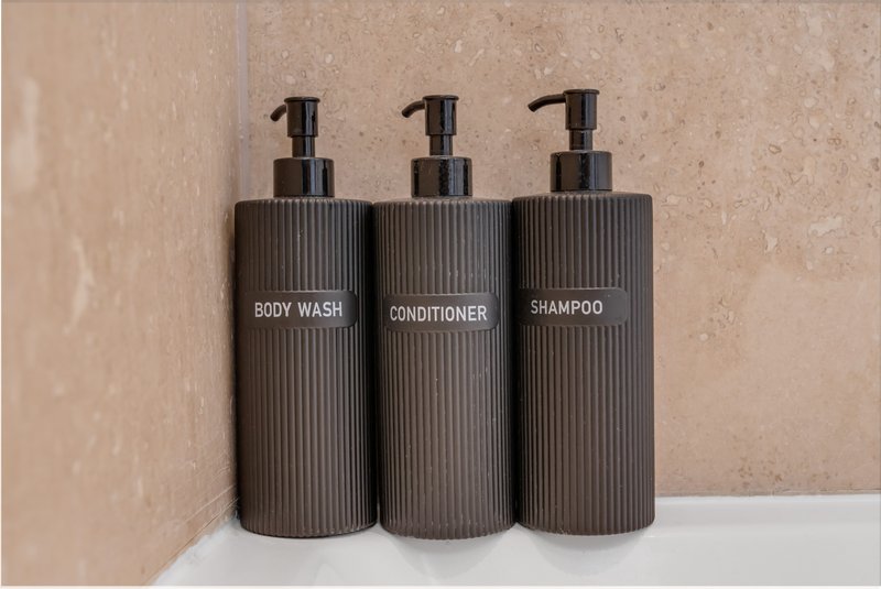 Three dark cylindrical bathroom dispensers labeled body wash, conditioner and shampoo on white shelf