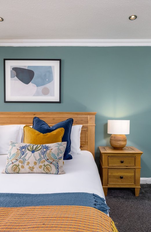 Bedroom with wooden bed frame, blue and yellow cushions, bedside table with lamp, and framed wall art