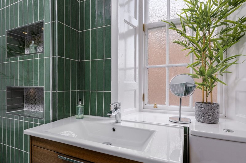 Modern bathroom with green tiled wall, white sink, wooden vanity, and window with plant
