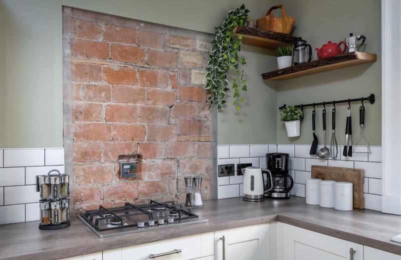 Kitchen corner with exposed brick backsplash, gas stovetop, white subway tiles, and wooden shelves with plants and kitchenware