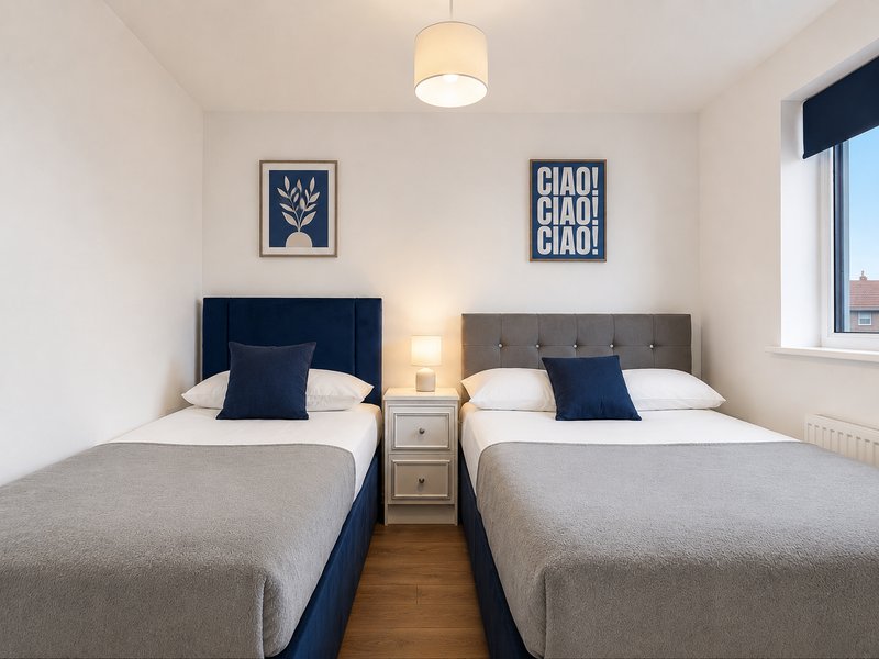 Twin bedroom with one single and one double bed, navy and grey headboards, bedside lamp and wall art and window