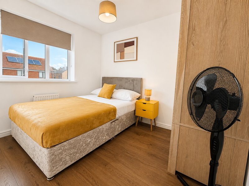 Modern bedroom with double bed, yellow bedding, wooden flooring and standing fan