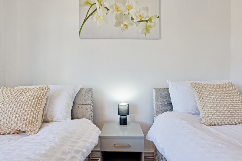Twin beds with white bedding, cream and grey cushions, small bedside table with lamp and framed floral artwork above