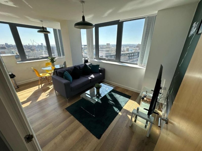 Modern apartment living room with black sofa, large windows overlooking city skyline, wooden flooring