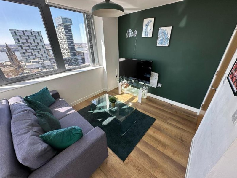 Modern living room with grey sofa, green accent wall, glass coffee table, and city views from large windows