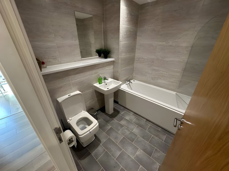 Modern bathroom with toilet, pedestal sink, bathtub, large mirror and grey tiling