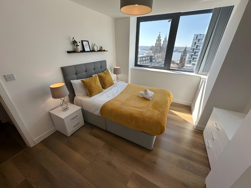 Bright modern bedroom with double bed, grey headboard, yellow cushions and throw, wooden flooring, city views from large window