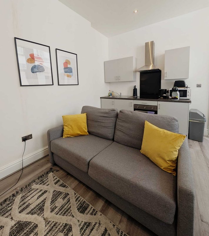 Studio living area with gray sofa, yellow cushions, kitchenette, and framed artwork