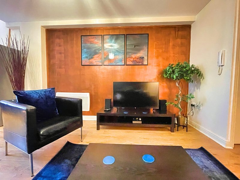 Modern living room with orange accent wall, black leather armchair, TV stand, and blue cushions
