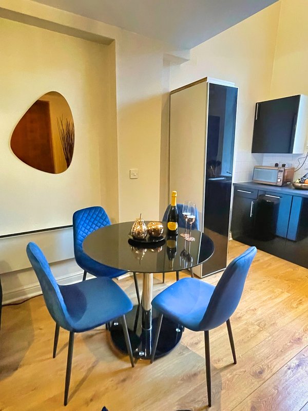 Modern dining area with round black table, blue chairs, wine bottles and kitchenette in background