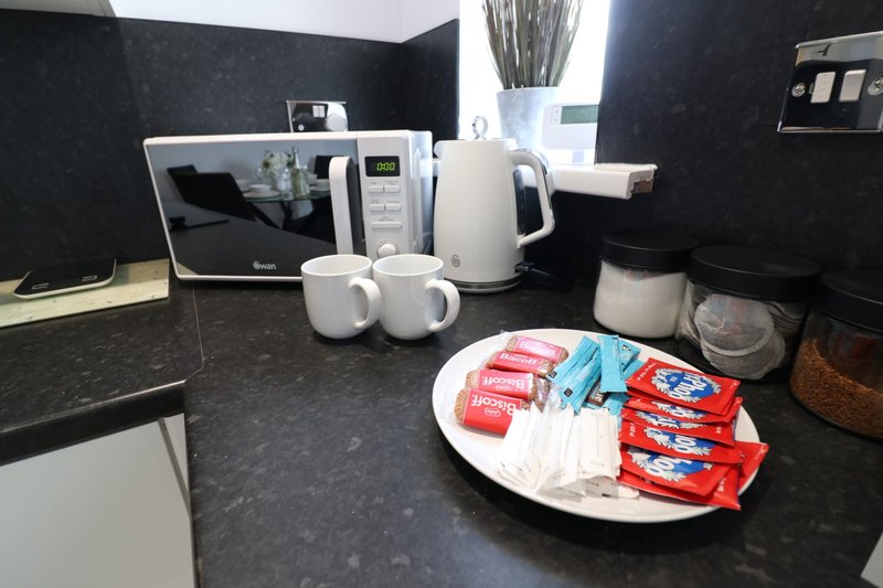 Modern kitchen with black countertop, microwave, kettle, mugs and welcome amenities on plate