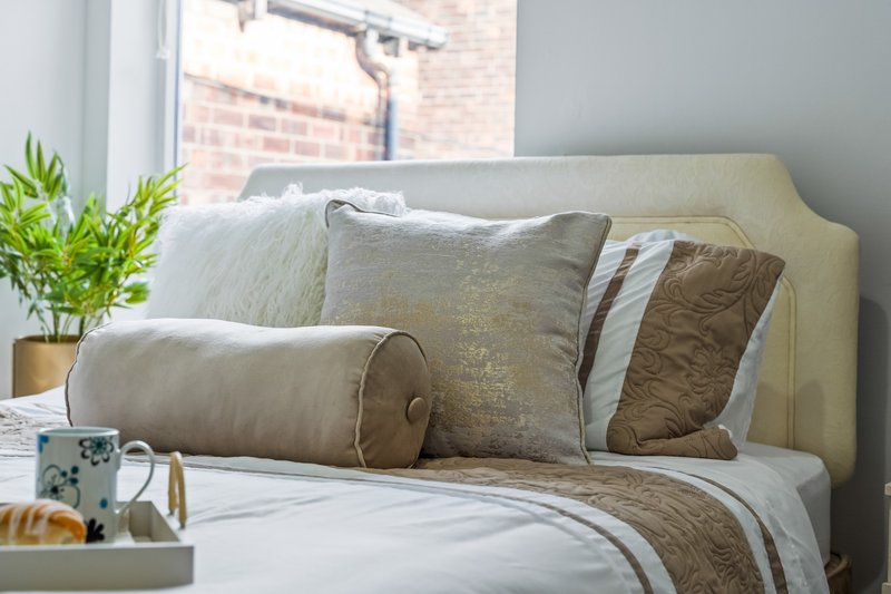 Bright bedroom with cream upholstered headboard, layered pillows and cushions, potted plant and mug on nightstand