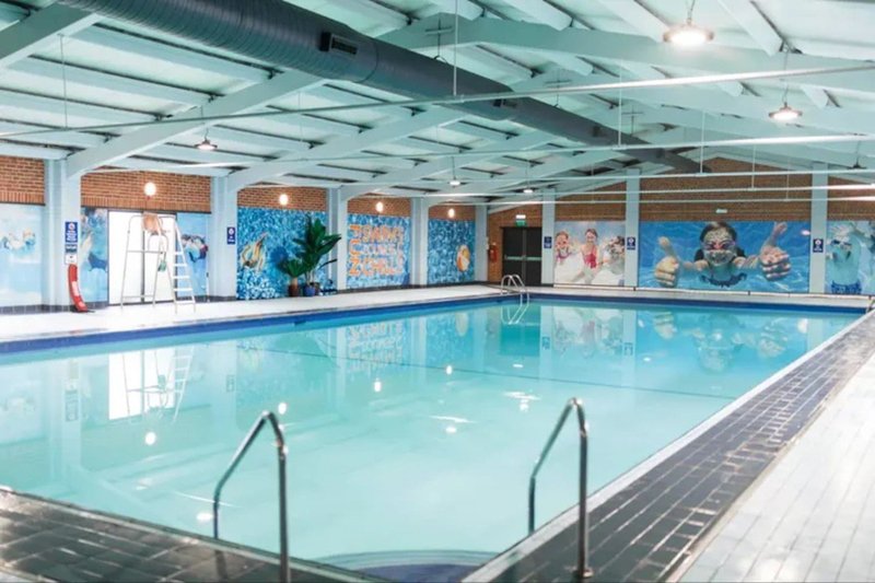 Indoor swimming pool with blue water, handrails, wooden decking, and glass-walled facility with natural light
