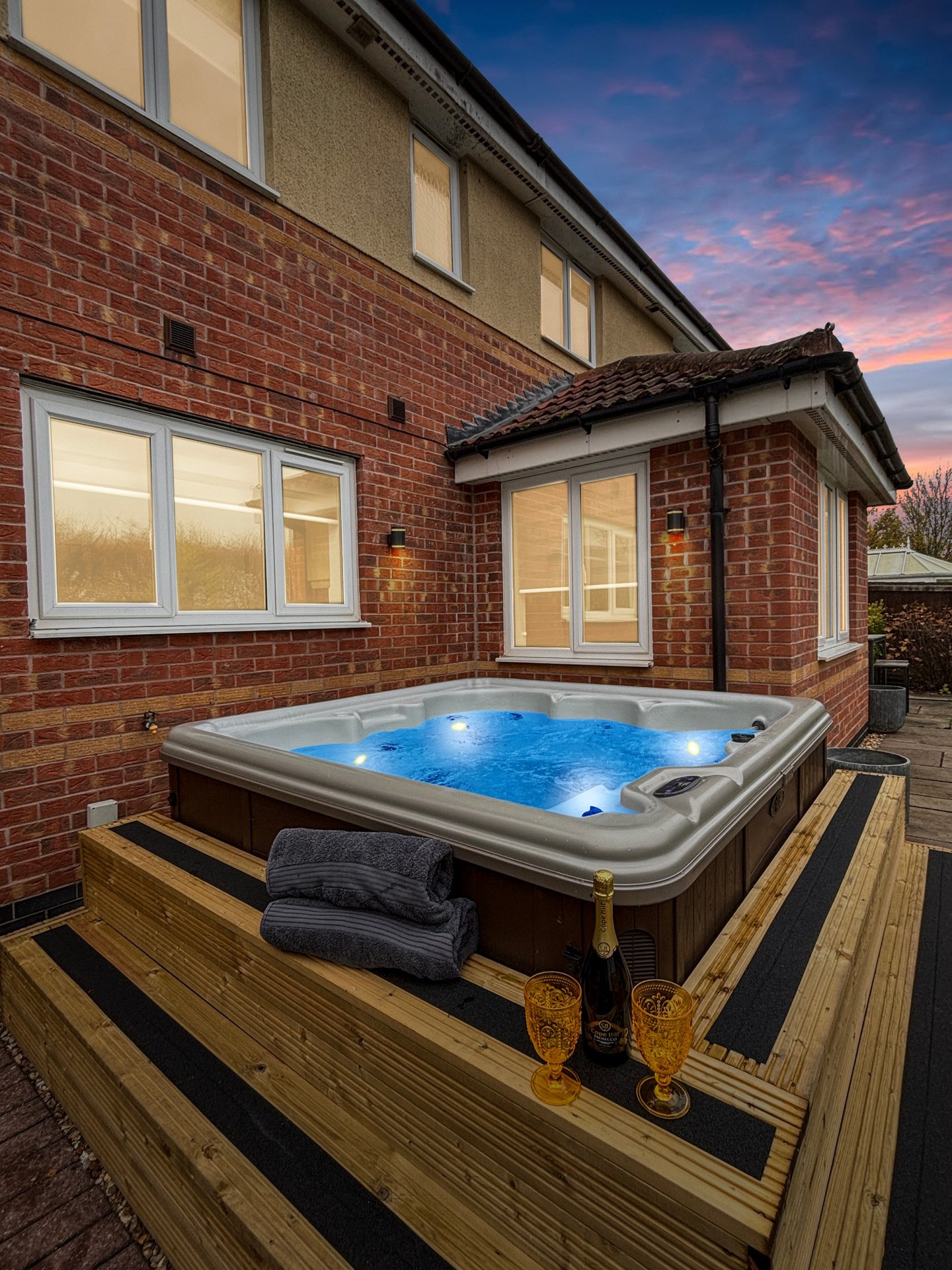 Hot tub on wooden deck at dusk with brick house and illuminated windows behind