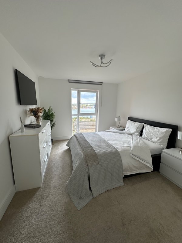 Modern bedroom with double bed, white dresser, wall-mounted TV, and window with coastal view