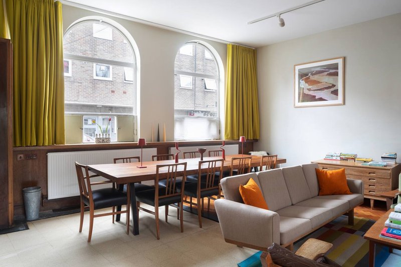 Open-plan living and dining room with sofa, wooden dining table, large arched windows, and mustard curtains