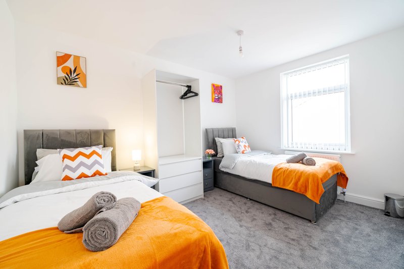 Bright twin bedroom with two single beds, orange and white bedding, grey upholstered headboards, window with blinds