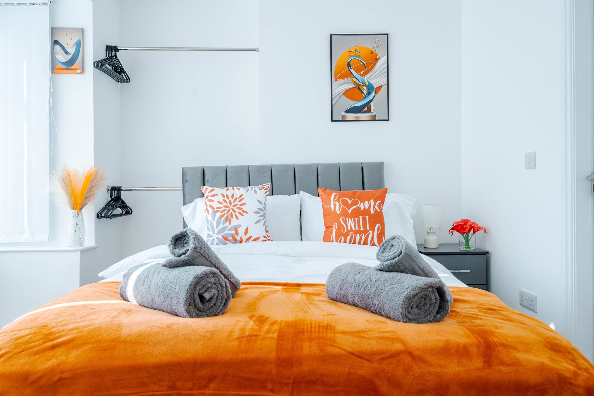 Modern bedroom with orange bedding, gray upholstered headboard, rolled towels, and decorative pillows