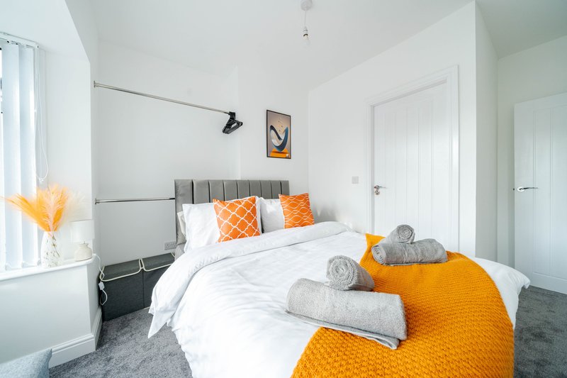 Modern bedroom with grey upholstered bed, white bedding, orange throw and cushions, rolled towels