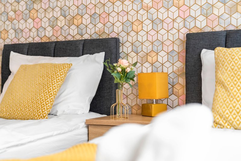 Bedroom with two beds, yellow cushions, white bedding, geometric wallpaper, wooden nightstand