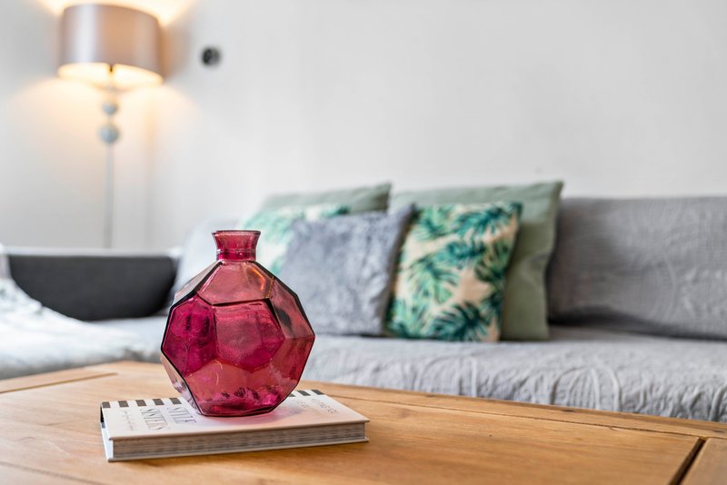 Living room with wooden table, pink decorative bottle, books, sofa with patterned cushions and ceiling lamp