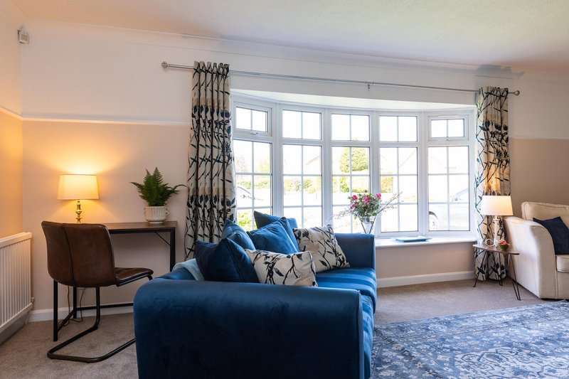 Bright living room with blue sofa, patterned cushions, desk lamp, and large bay windows with curtains