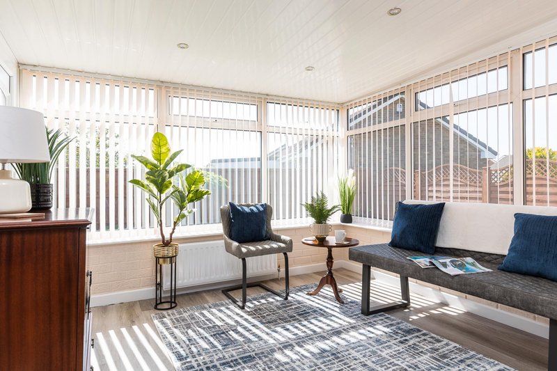 Bright conservatory with vertical blinds, potted plants, seating area and wooden furniture
