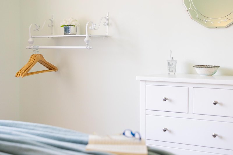 Bright bedroom corner with white dresser, wall shelf, wooden hangers and folded linens