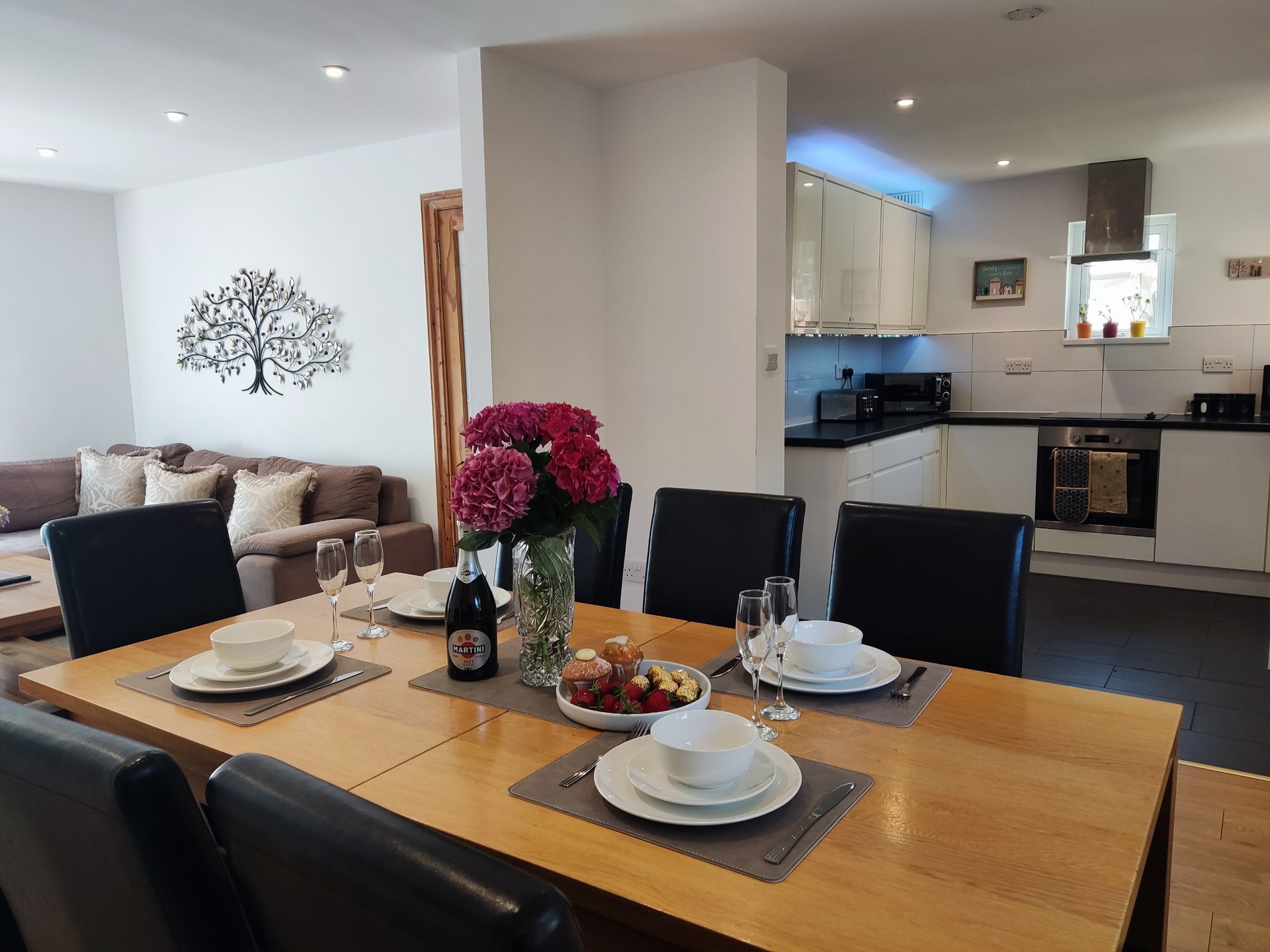 Open-plan living and kitchen with wooden dining table set for two, modern cabinetry, and tree artwork