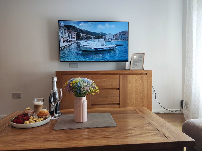 Living room with wooden table, wall-mounted TV displaying coastal scene, and wooden sideboard
