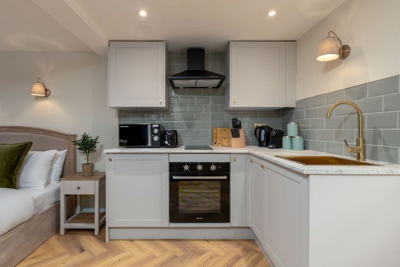 Modern studio kitchen with white cabinetry, black range hood, and grey tile backsplash beside bed